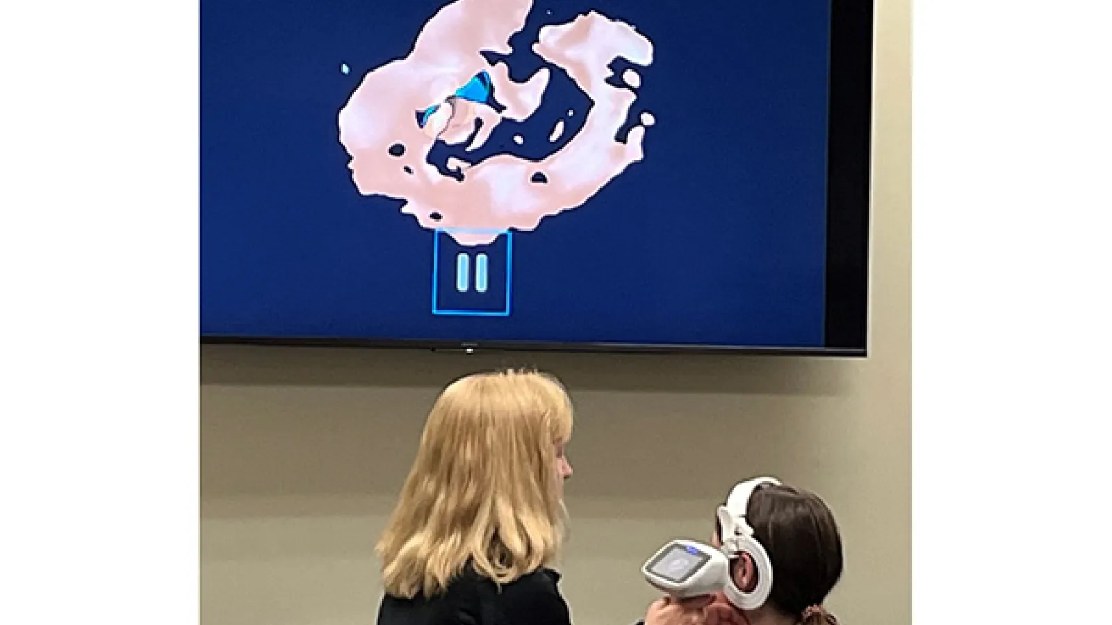 Audiologist holds equipment while patient wears device on head in front of large monitor with scan results