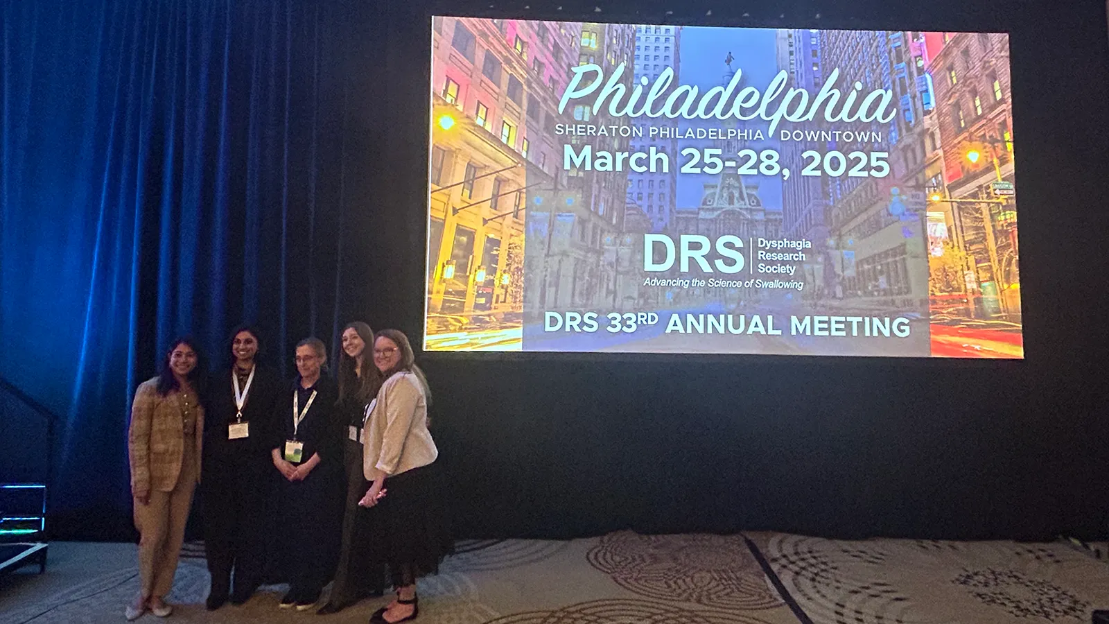 Five professionals posing in front of screen projection: "Philadelphia - DRS 33rd Annual Meeting"