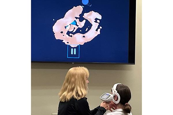 Audiologist holds equipment while patient wears device on head in front of large monitor with scan results
